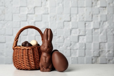 Photo of Wicker basket with sweet chocolate Easter eggs and bunny on table against light background, space for text