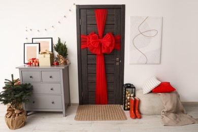 Wooden door with beautiful red bow near chest of drawers, evergreen trees and gift boxes indoors. Christmas decoration