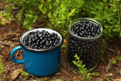 Jar and cup of delicious bilberries on ground in forest