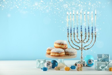 Image of Hanukkah celebration. Menorah with burning candles, dreidels, gift boxes and donuts on white wooden table against light blue background, space for text
