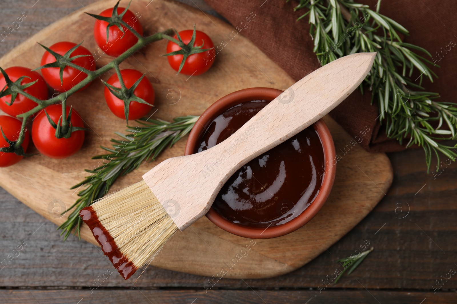 Photo of Marinade in bowl, basting brush, tomatoes and rosemary on wooden table, flat lay