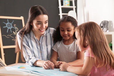 Photo of Cute little children with nursery teacher reading book at table in kindergarten. Indoor activity