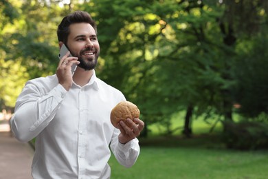 Lunch time. Young businessman with hamburger talking on smartphone in park, space for text