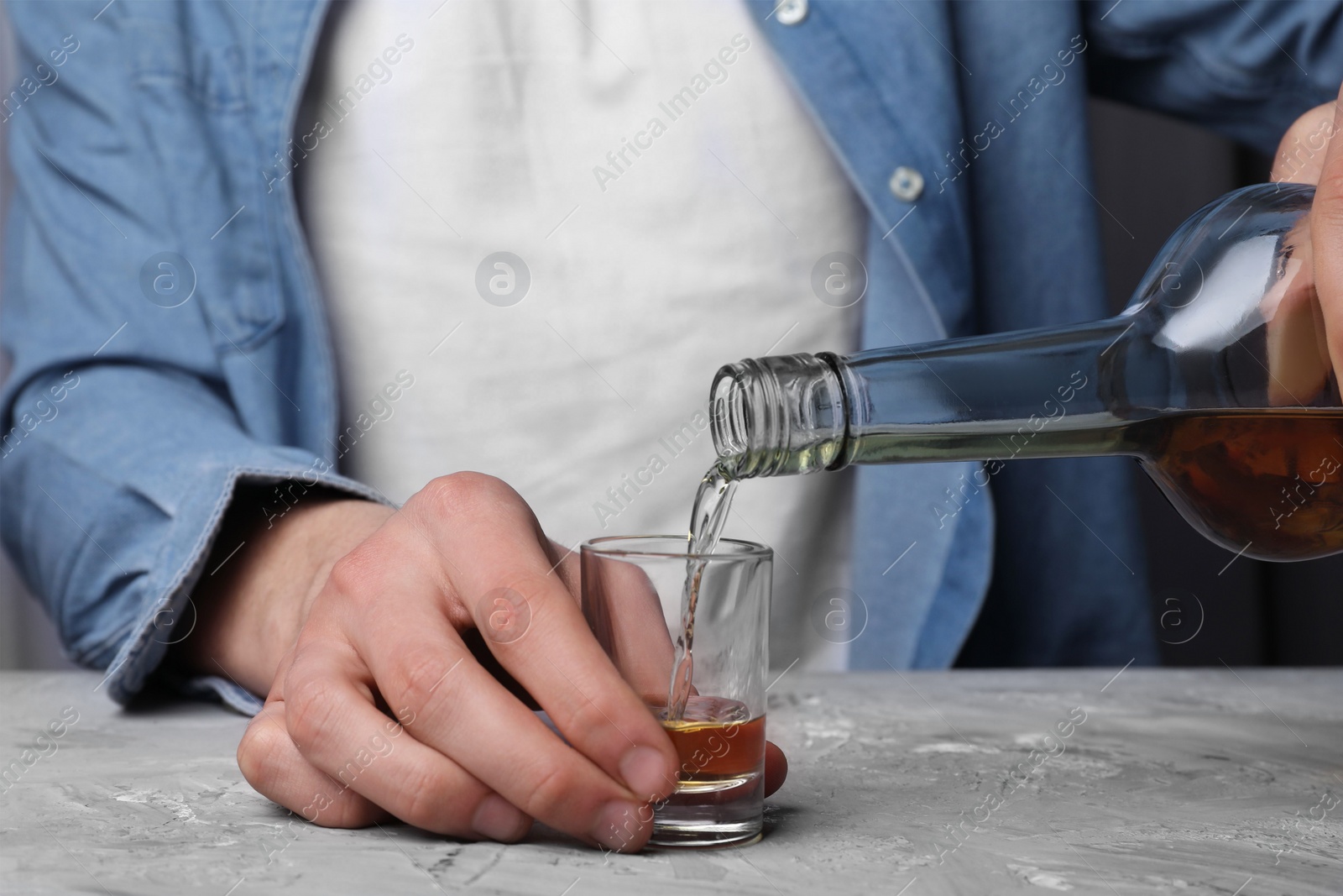 Photo of Alcohol addiction. Man pouring whiskey from bottle into glass at grey textured table, closeup