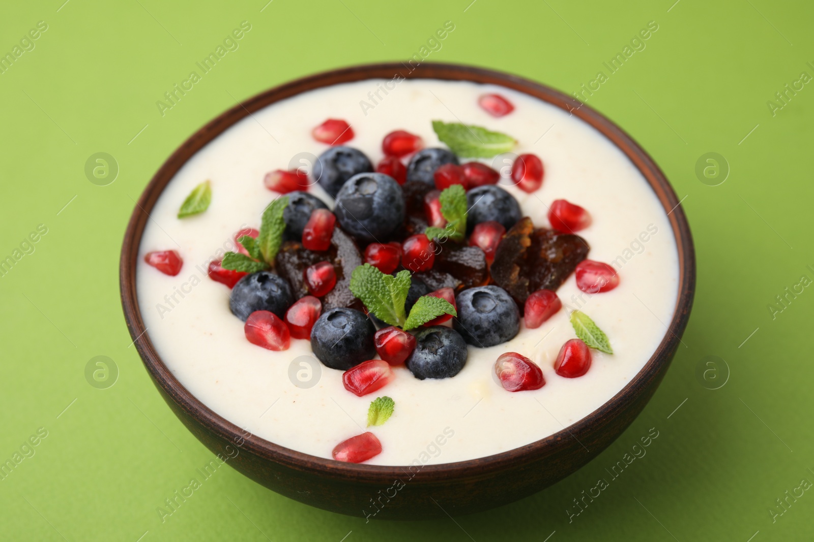 Photo of Delicious semolina pudding with blueberries, pomegranate, dates and mint in bowl on green table, closeup