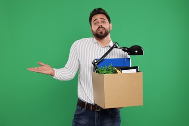 Confused unemployed man with box of personal office belongings on green background