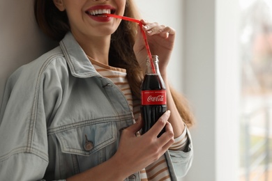 MYKOLAIV, UKRAINE - NOVEMBER 28, 2018: Young woman with bottle of Coca-Cola indoors, closeup. Space for text