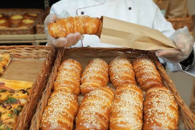 Photo of Baker putting fresh bun into paper bag over tray with pastry, closeup