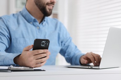 Photo of Young man using smartphone while working with laptop at white table in office, closeup