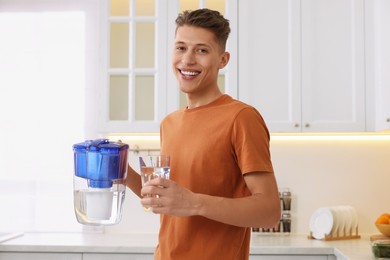 Happy man with filter jug and glass of clear water in kitchen