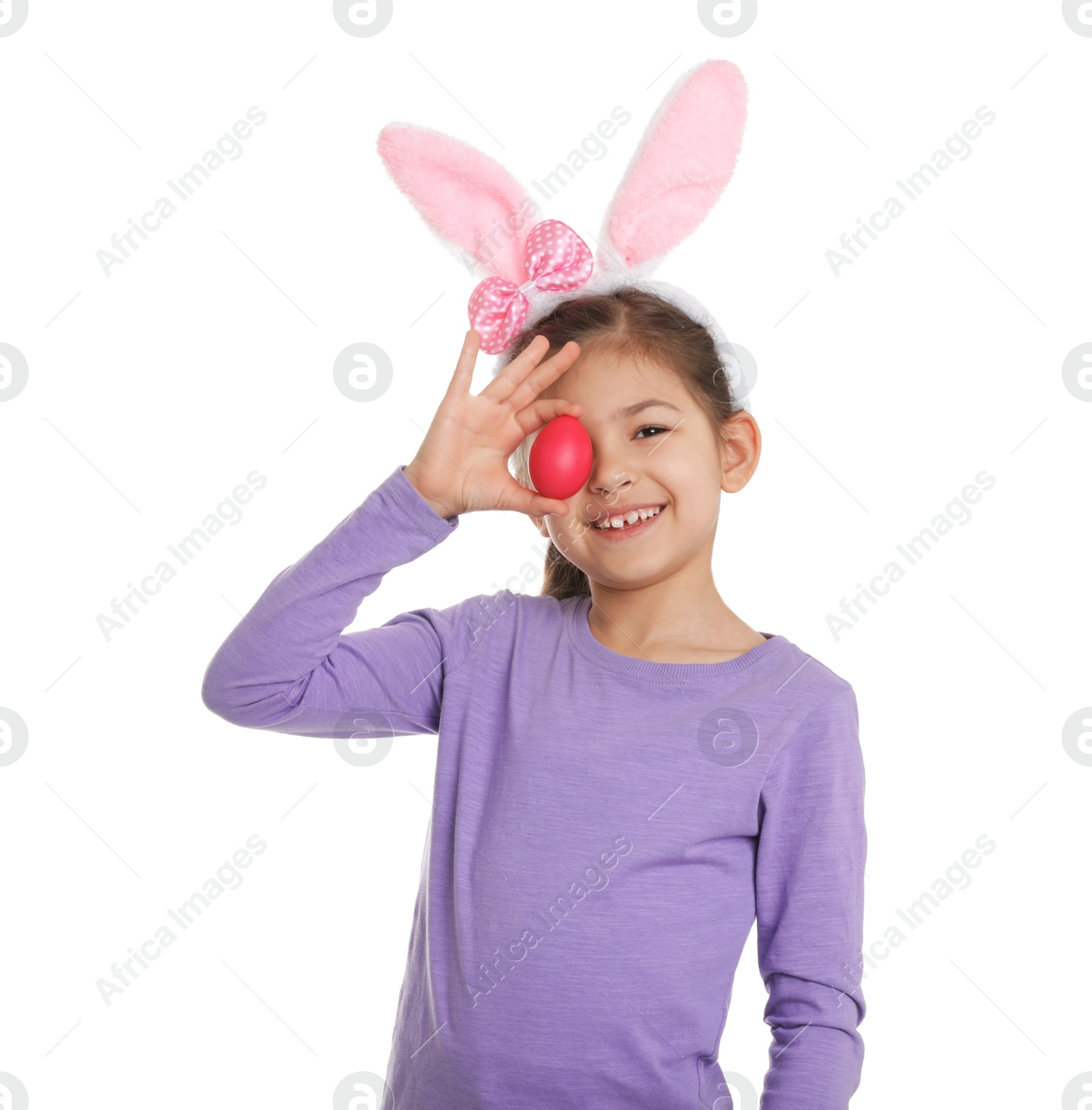 Photo of Little girl in bunny ears headband holding Easter egg near eye on white background