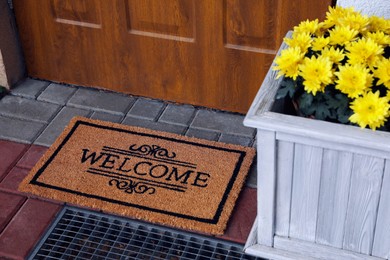 Doormat with word Welcome and beautiful flowers on floor near entrance