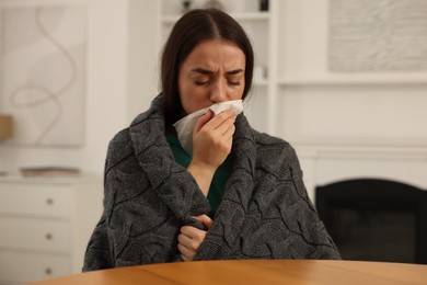 Photo of Sick woman wrapped in blanket with tissue blowing nose at wooden table indoors. Cold symptoms