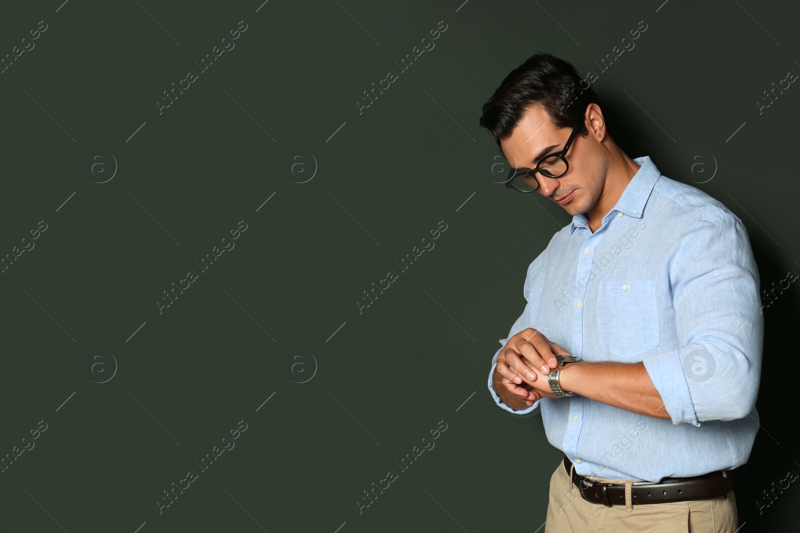 Photo of Handsome young man in stylish clothes with glasses checking time on dark background. Space for text