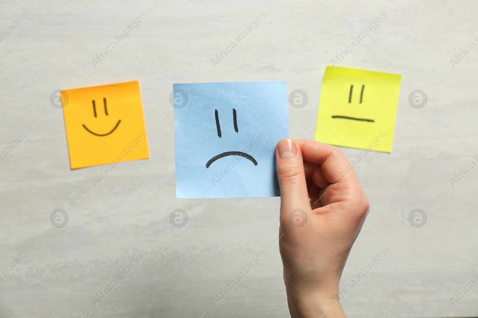 Photo of Woman holding sticker with sad face on light background, closeup