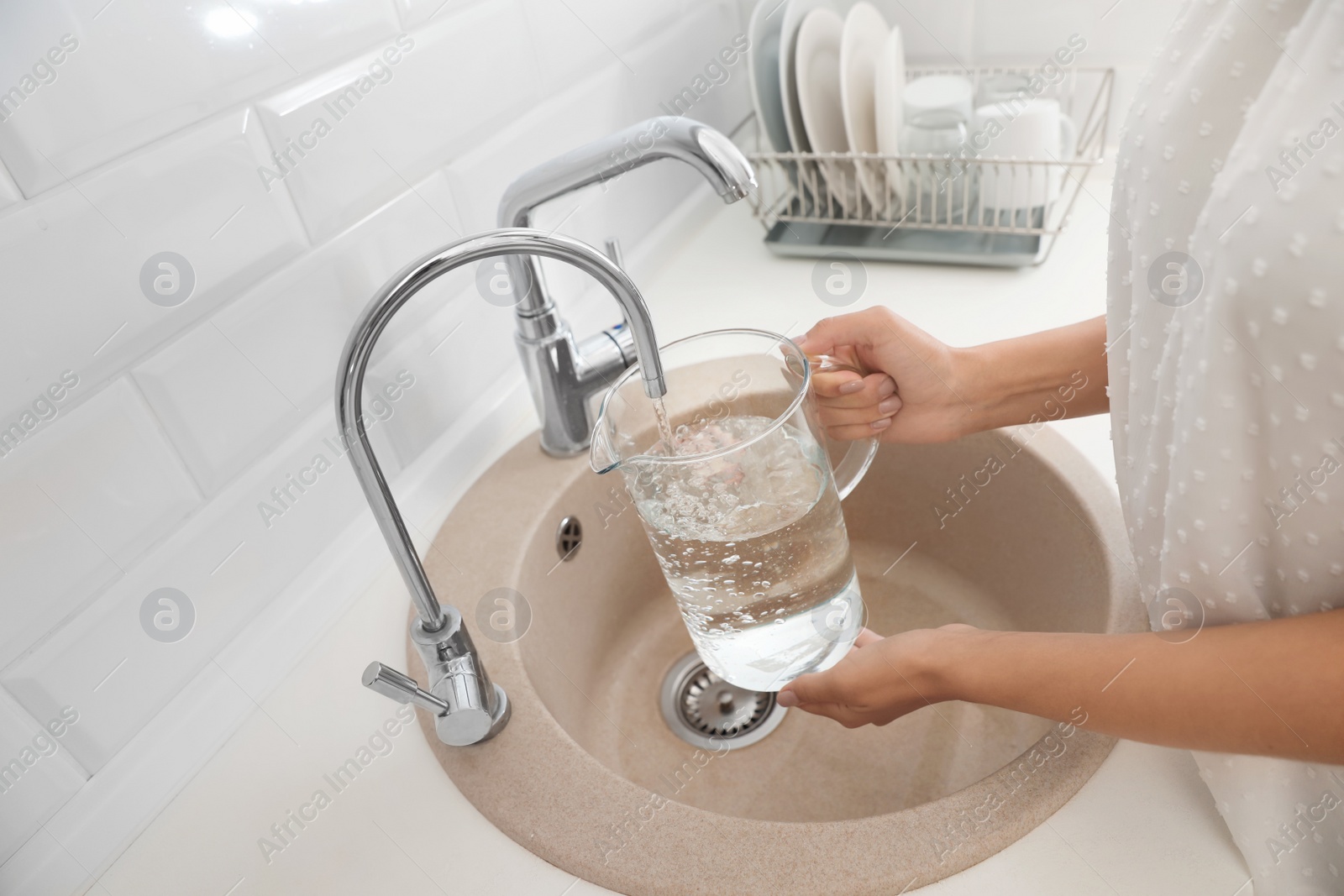 Photo of Woman pouring water into glass jug in kitchen, closeup