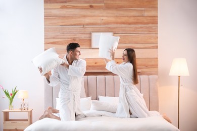Happy couple in bathrobes having pillow fight on bed at home