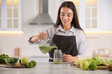 Young woman pouring fresh green juice into mason jar at table in kitchen