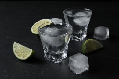 Photo of Shot glasses of vodka with lime slices and ice on black table