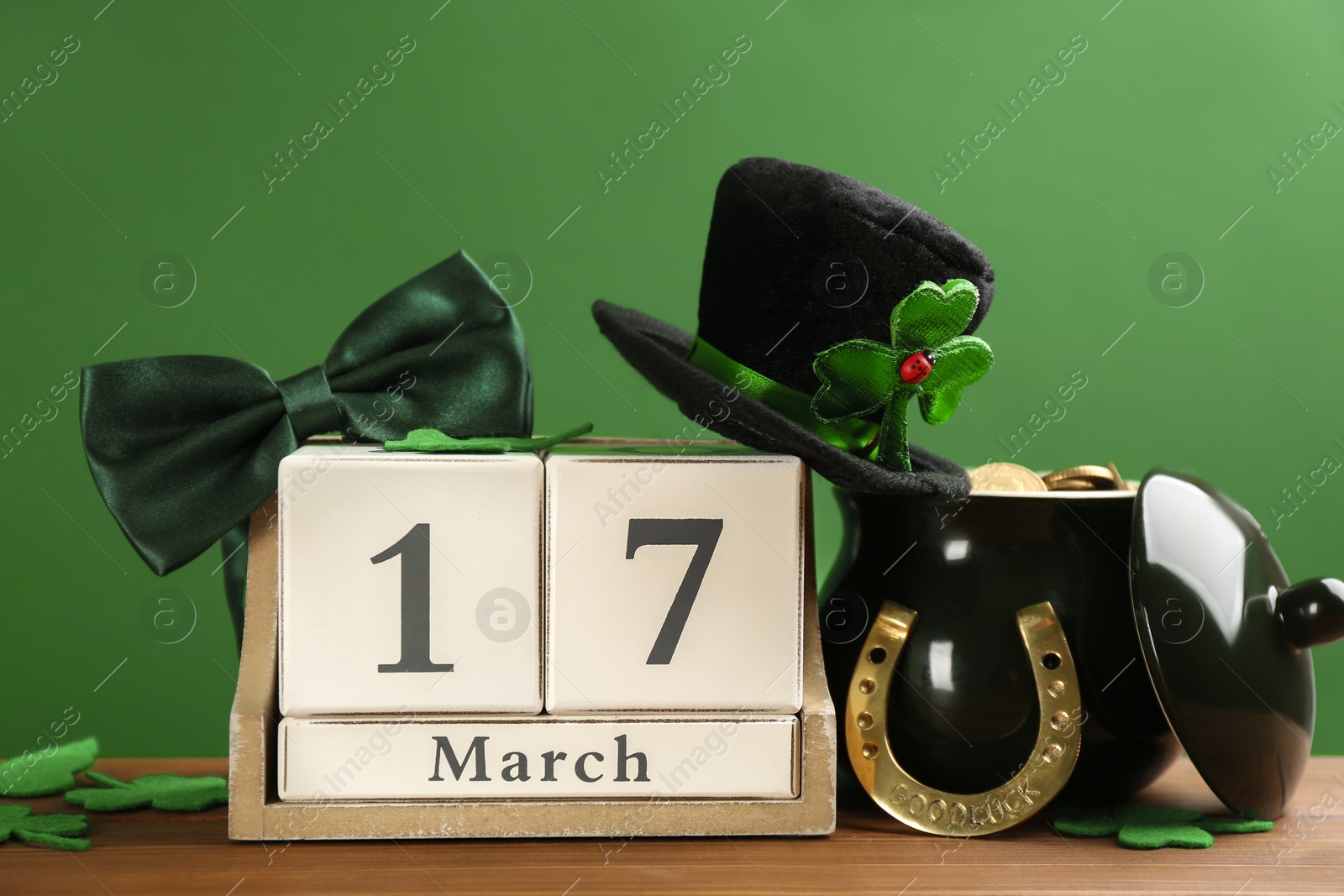 Photo of Composition with leprechaun hat, block calendar and pot of gold on wooden table against green background. St Patrick's Day celebration
