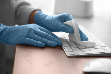 Photo of Woman in latex gloves cleaning computer keyboard with wet wipe at table, closeup