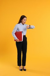 Shocked woman with folder looking at her watch on orange background