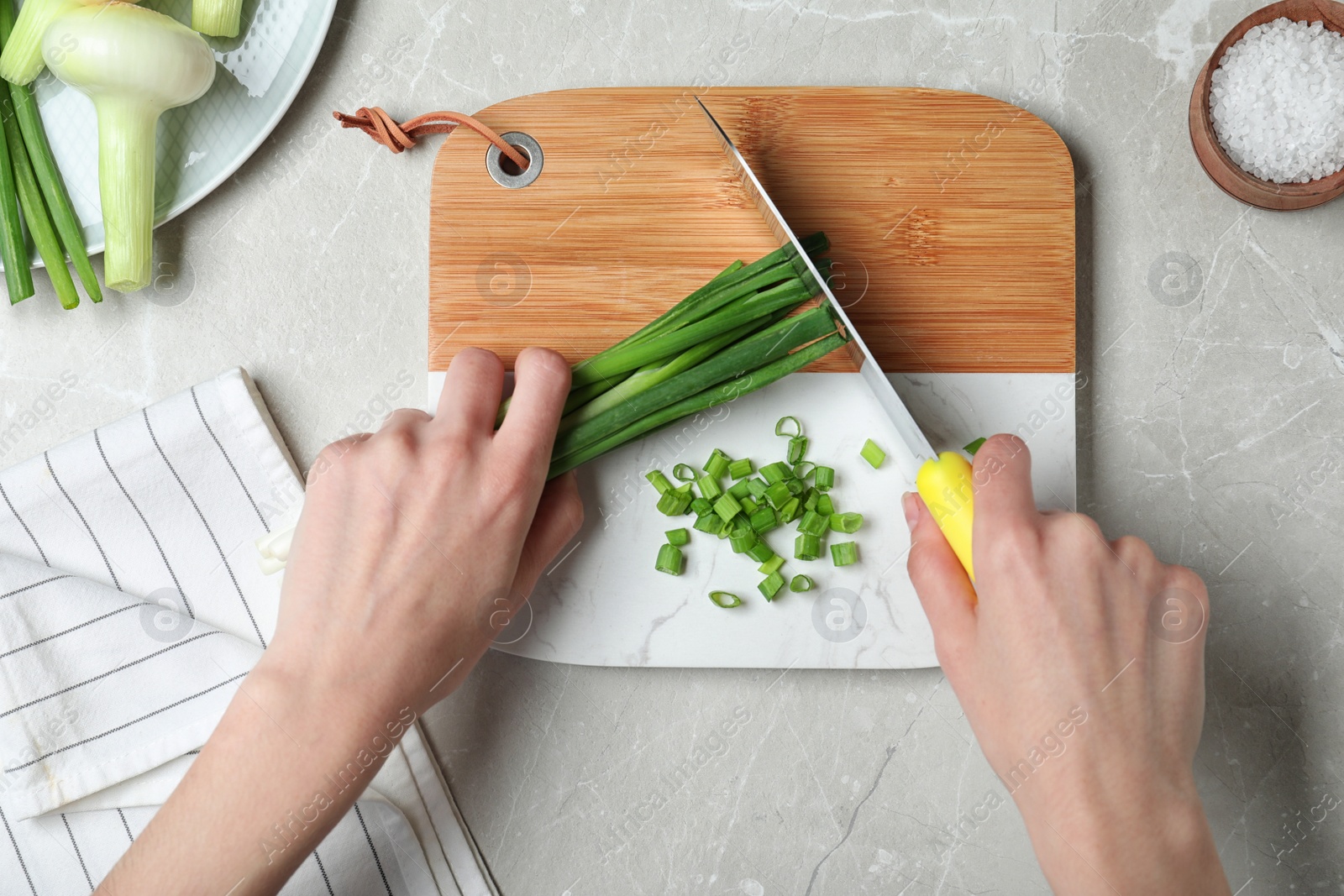 Photo of Woman cutting fresh green onion on wooden board at marble table, top view