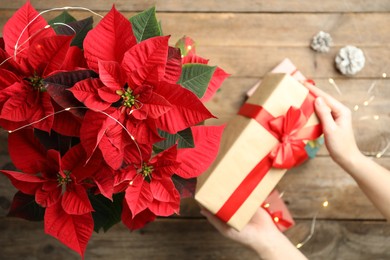 Photo of Woman with gift near poinsettia (traditional Christmas flower) at wooden table, top view