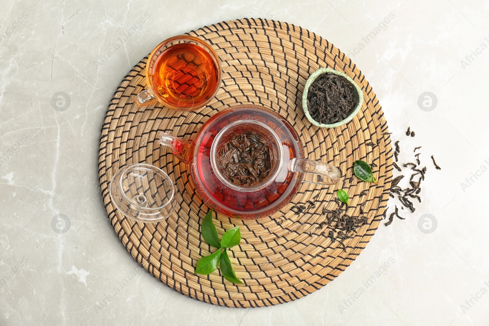 Photo of Teapot with hot aromatic tea, dry and fresh leaves on wicker mat, top view