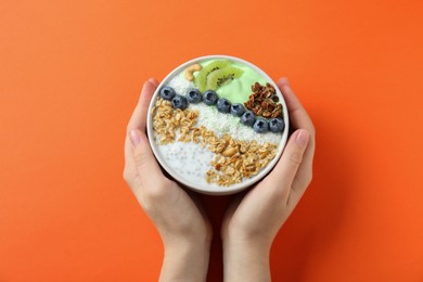 Woman holding tasty smoothie bowl with fresh kiwi fruit, blueberries and oatmeal at orange table, top view