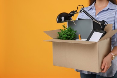 Photo of Unemployed woman holding box with personal office belongings on orange background, closeup. Space for text