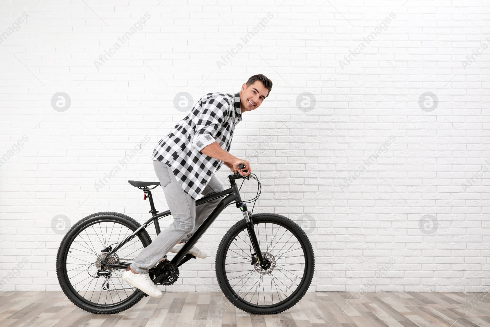 Photo of Handsome young man with modern bicycle near white brick wall indoors