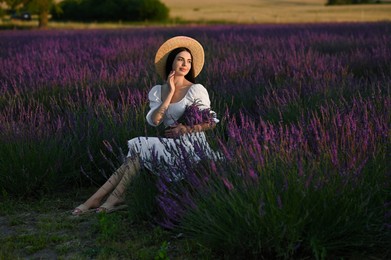 Beautiful young woman with bouquet sitting in lavender field at sunset