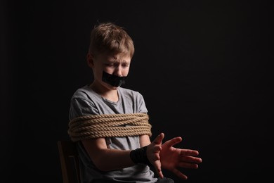 Photo of Little boy with taped mouth tied up and taken hostage against dark background. Space for text