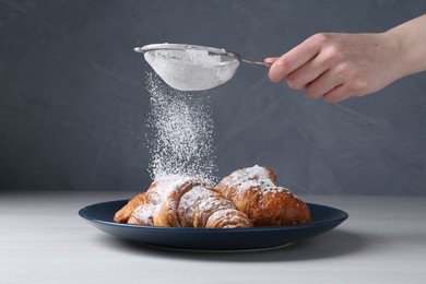 Woman with sieve sprinkling powdered sugar onto croissants at white wooden table, closeup