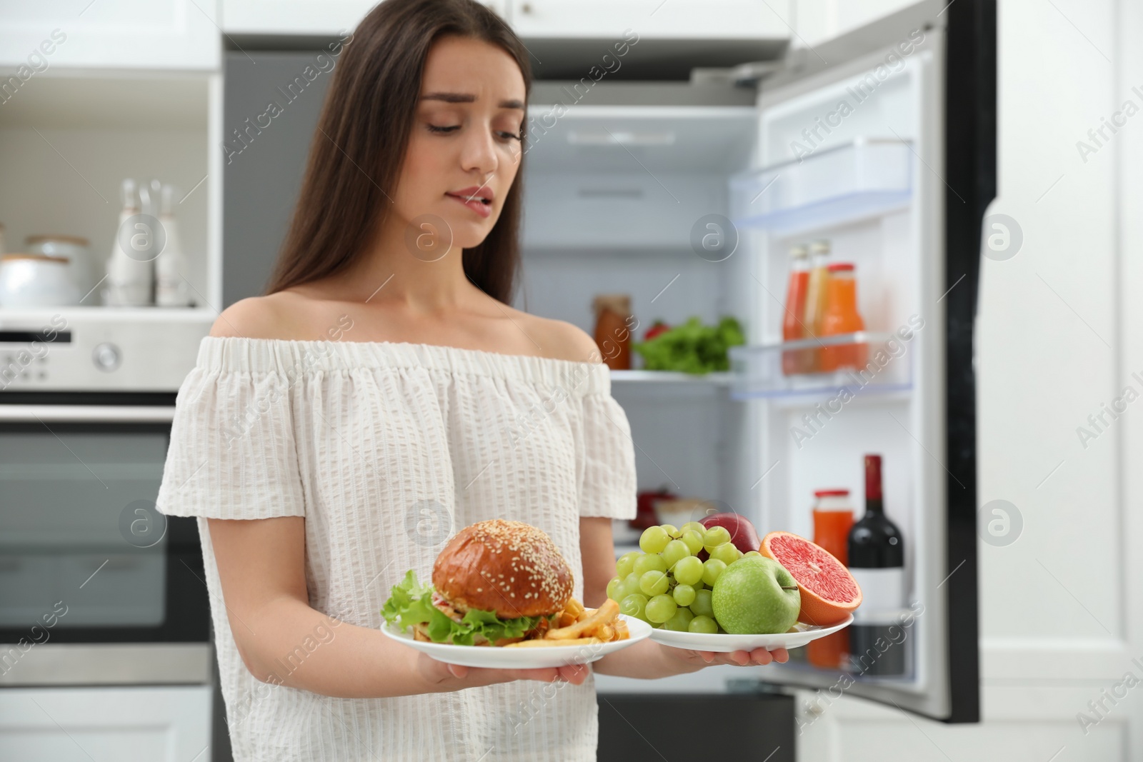 Photo of Woman choosing between fruits and burger with French fries near refrigerator in kitchen