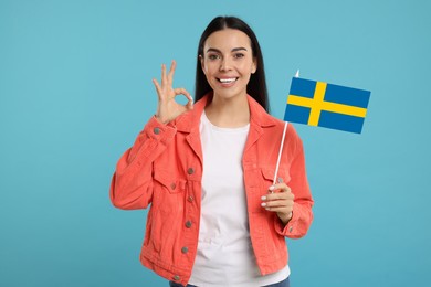 Happy young woman with flag of Sweden showing OK gesture on light blue background