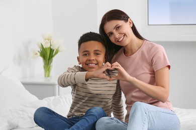 Photo of Mother and her African American son making heart with hands on bed at home. International family