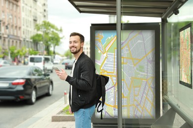 Image of Young man with smartphone and backpack waiting for public transport at bus stop