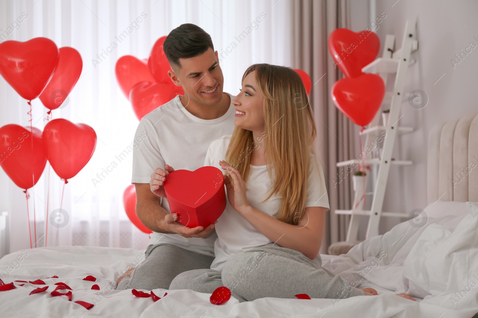 Photo of Man presenting gift to his girlfriend in room decorated with heart shaped balloons. Valentine's day celebration