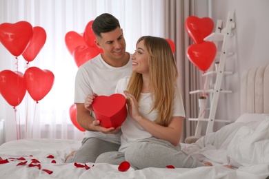 Man presenting gift to his girlfriend in room decorated with heart shaped balloons. Valentine's day celebration