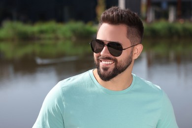 Handsome smiling man in sunglasses near river, space for text