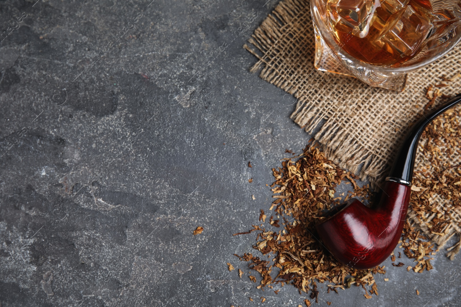 Photo of Smoking pipe with tobacco and glass of whiskey on grey table, flat lay. Space for text