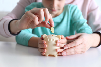 Photo of Mother and son putting coin into piggy bank at table indoors, closeup