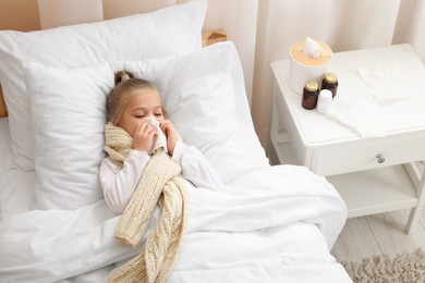 Photo of Sick girl with scarf and tissue lying in bed while blowing nose indoors, above view. Cold symptoms