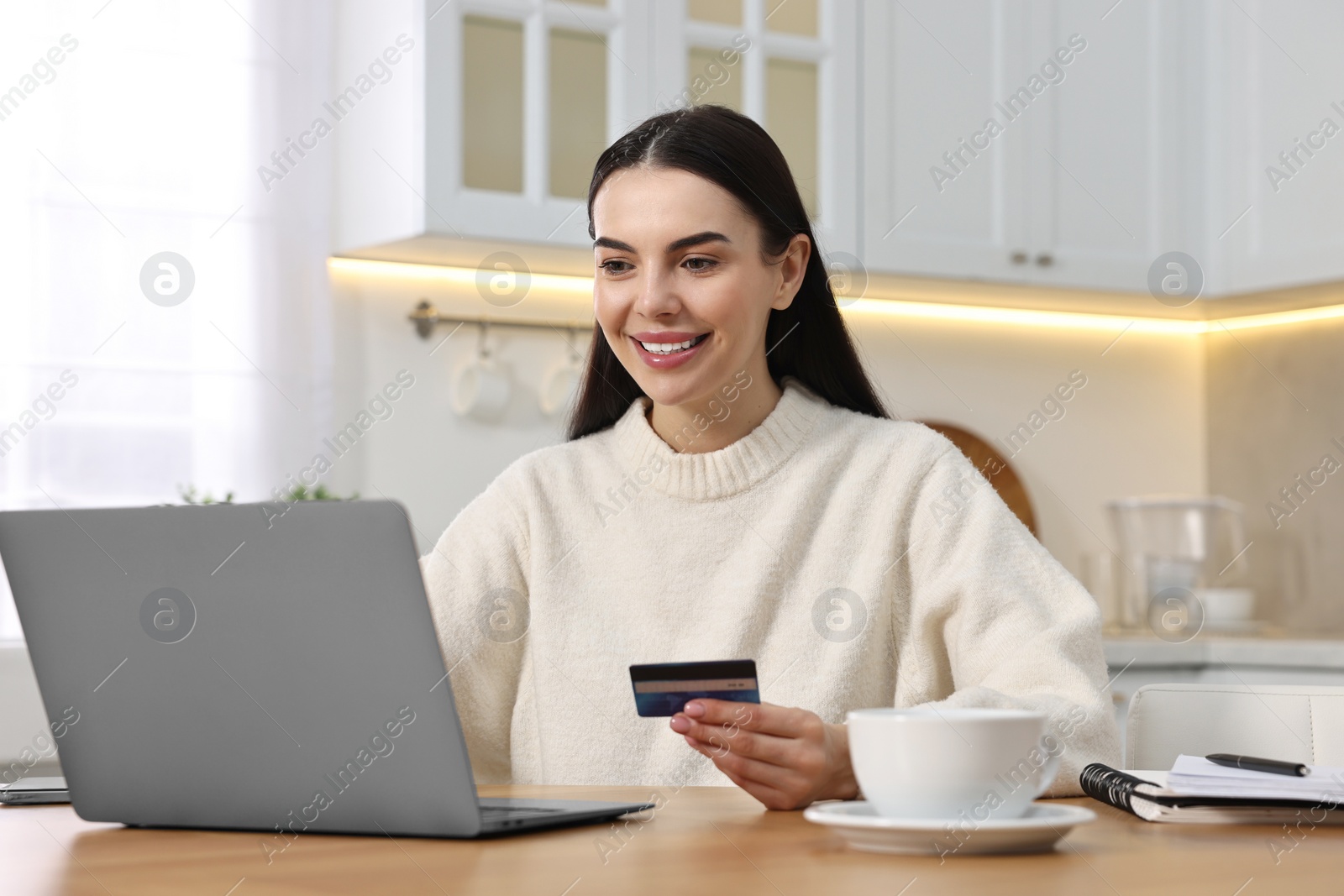 Photo of Happy young woman with credit card using laptop for shopping online at wooden table in kitchen