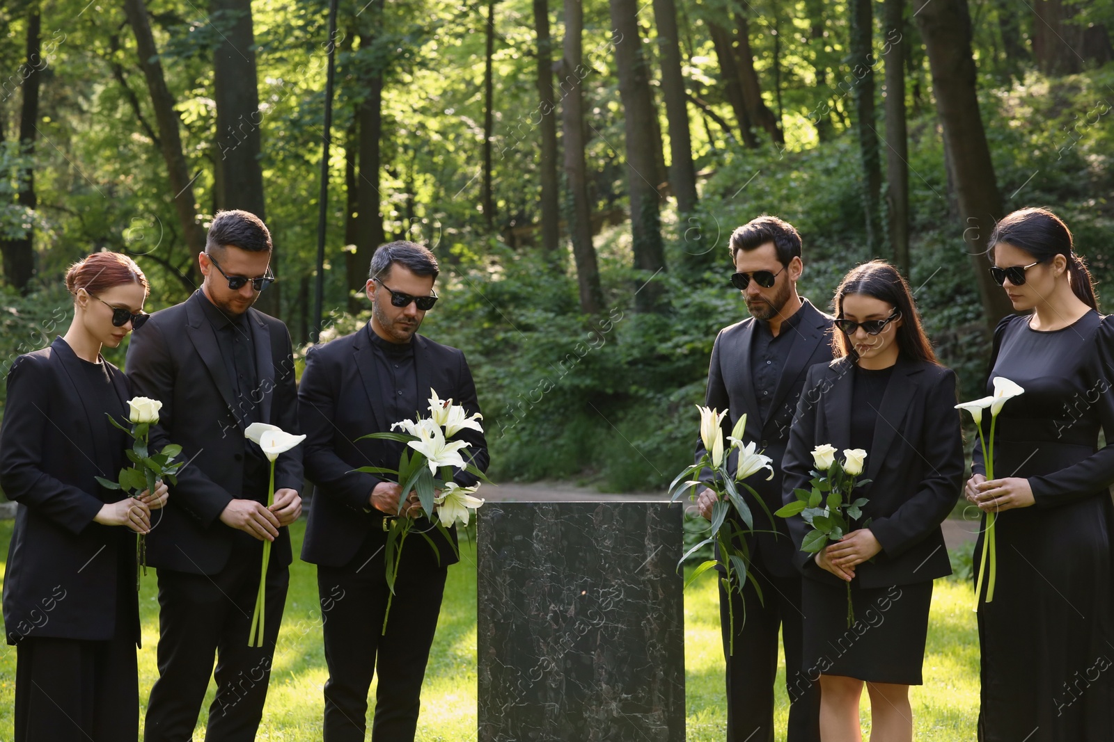 Photo of Sad people with flowers mourning near granite tombstone at cemetery outdoors. Funeral ceremony