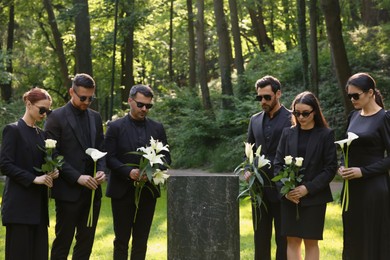 Sad people with flowers mourning near granite tombstone at cemetery outdoors. Funeral ceremony