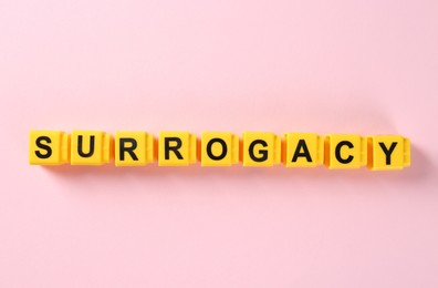 Photo of Word Surrogacy made of yellow cubes on pink background, flat lay