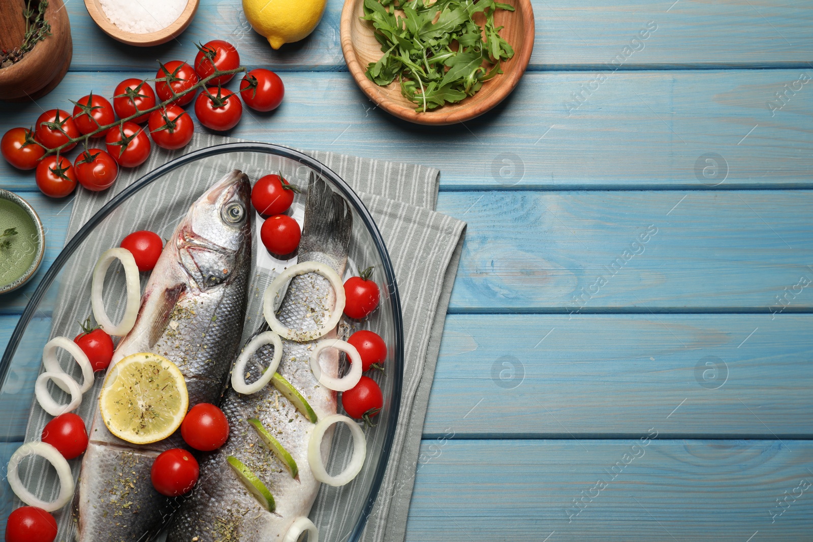 Photo of Glass baking tray with sea bass fish and ingredients on light blue wooden table, flat lay. Space for text